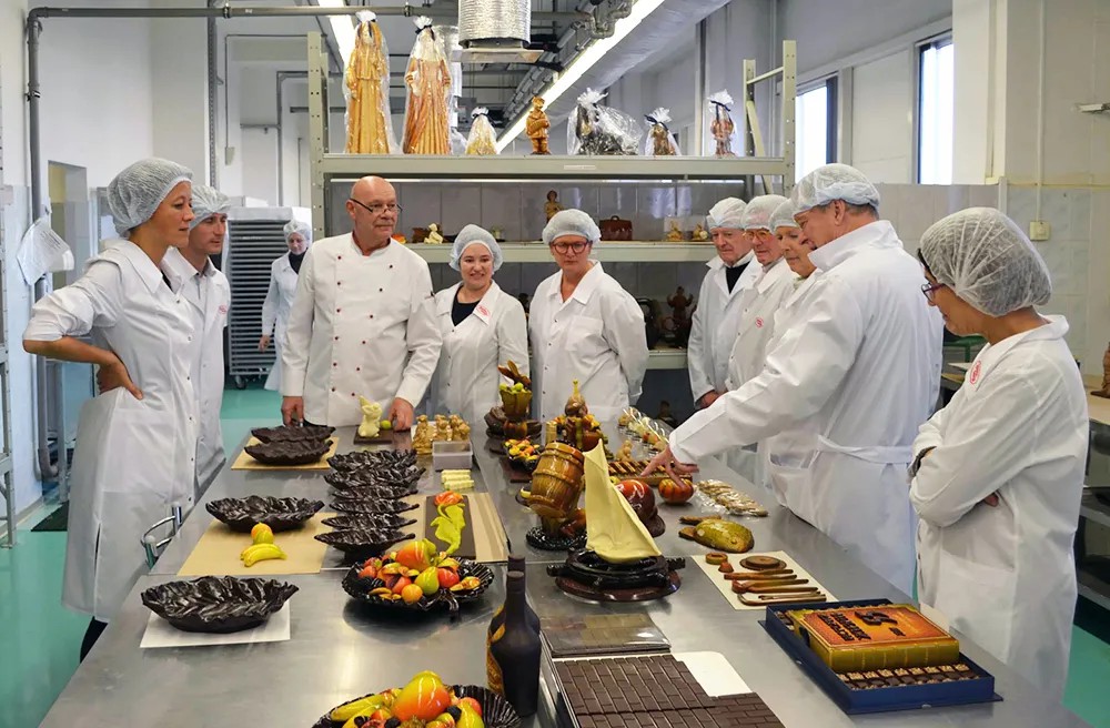 Фотография из цеха ручной работы: мастер-шоколатье Владимир Ефимович Коннэ проводит экскурсию по цеху для потомков легендарных немецких кондитеров Эйнема и Гейса