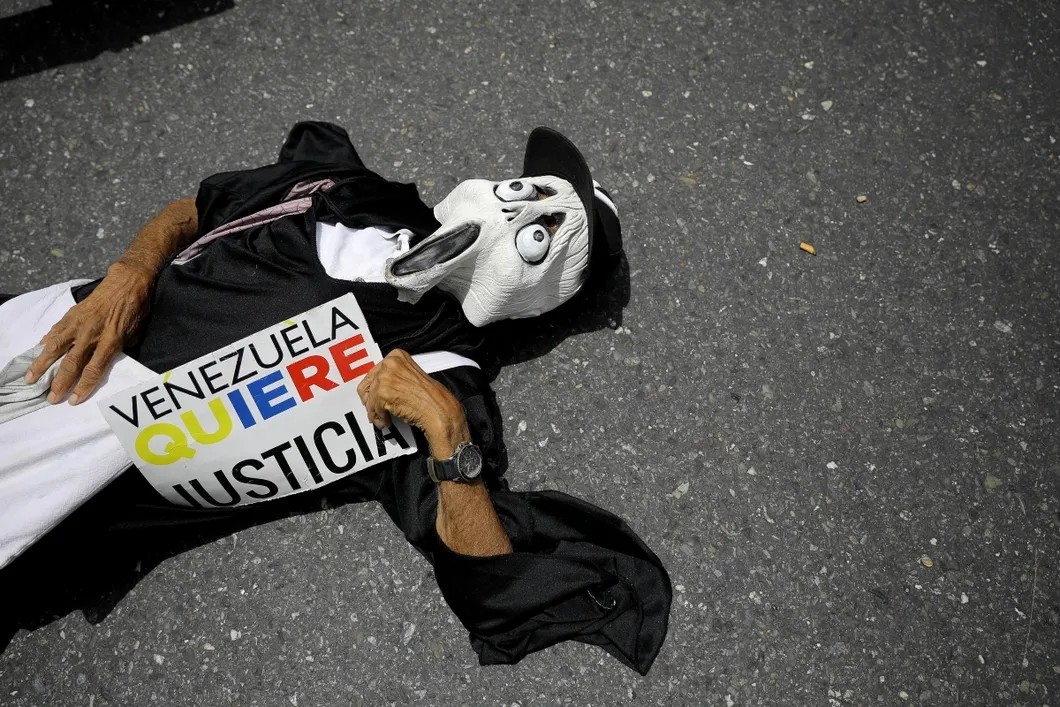 Один из протестующих в маске и плакатом со словами «Венесуэла хочет справедливости» в руках лежит на земле. Фото: Reuters