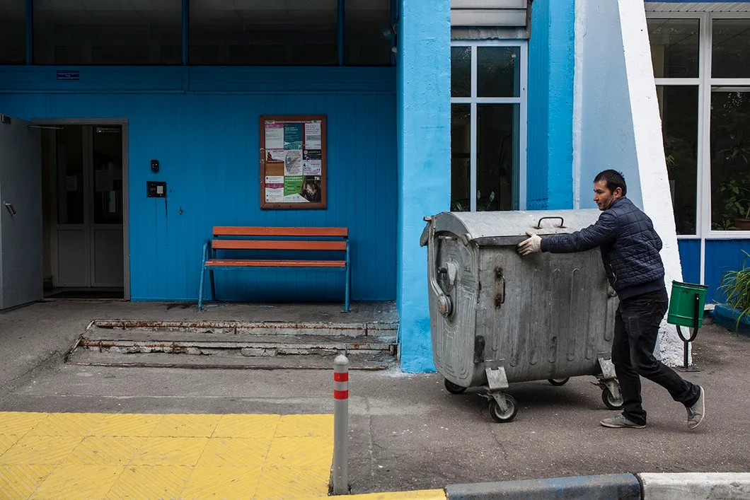 Мухиддин катит мусорный контейнер у подъезда дома, территорию которого он убирает. Фото: Влад Докшин / «Новая»